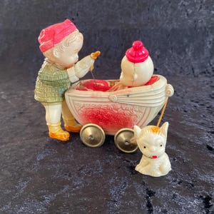 May include: A vintage toy set featuring a child figure in a green coat and pink hat, tending to a baby in a white carriage with red accents. A small white dog with orange markings is also present. The toy has gold-colored wheels.