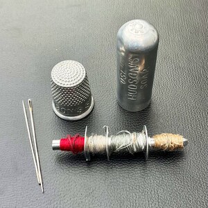 May include: A silver thimble, a silver spool of thread with red, beige, and gray thread, two silver needles, and a silver container with the text "SOUS HUDSON'S 25D" on it.