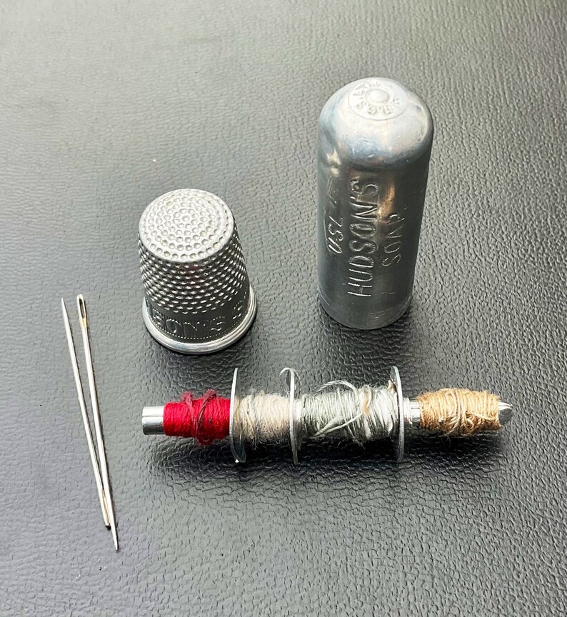 May include: A silver thimble, a silver spool of thread with red, beige, and gray thread, two silver needles, and a silver container with the text "SOUS HUDSON'S 25D" on it.