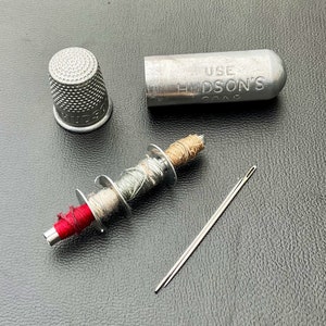 May include: A silver thimble, a small spool of thread with three different colors of thread, a needle, and a silver container with the text "USE HUDSON'S SOAP" on it.