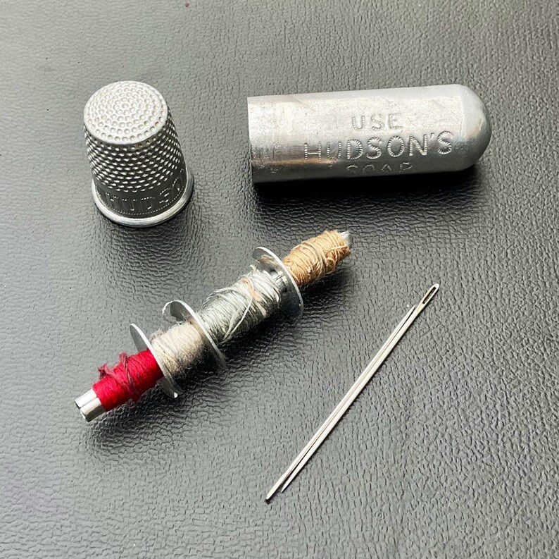 May include: A silver thimble, a small spool of thread with three different colors of thread, a needle, and a silver container with the text "USE HUDSON'S SOAP" on it.