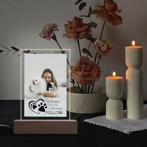 May include: A rectangular, illuminated photo frame with a wooden base. The frame displays a photo of a person with two dogs, and the text "Wherever you go I will always FOLLOW" with paw prints. Candles and flowers are in the background.