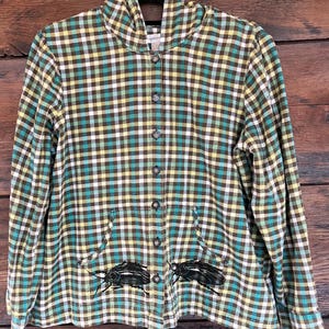 May include: A button-up hooded shirt with a plaid pattern in shades of green, yellow, brown, and white. The shirt features two front pockets with black insect embroidery. The shirt is hanging on a black hanger against a wooden background.