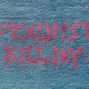 May include: Close-up of blue denim fabric with the words "FEMINIST KILLJOY" in a dripping, gothic-style red font. The text is centered and takes up most of the frame, with the denim texture visible.