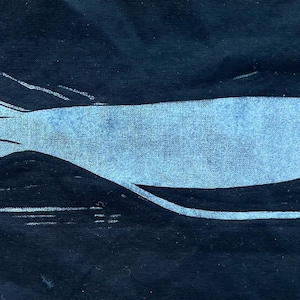 May include: A light blue linocut print of a fish on a dark blue background. The fish has a rounded body, a forked tail, and two small eyes. The linocut style gives the image a textured appearance, with visible lines and imperfections.