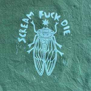 May include: Green fabric with a white insect graphic and the words "SCREAM FUCK DIE" in a circular arrangement. The insect has detailed wings and a star above its head.