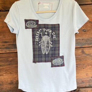 May include: A light blue short-sleeve t-shirt featuring a central rectangular dark plaid fabric patch with a white graphic of a cicada and the text "SCREAM FUCK DIE" printed above it, along with two smaller square plaid patches with spiral designs.