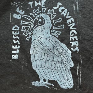 May include: A linocut print on black paper features a stylized owl with the words "Blessed Be" and "The Scavengers" in a light blue color. The owl is the central focus, with detailed feather patterns.