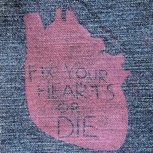 May include: Close-up of a denim fabric with a large, pink heart shape. The heart contains the handwritten text "FIX YOUR HEARTS OR DIE" in dark blue. The denim has a textured appearance.