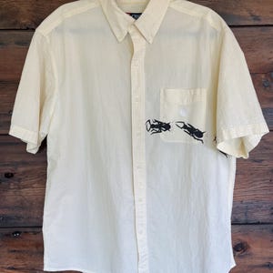 Può includere: Una camicia a maniche corte color giallo chiaro con bottoni e un sottile motivo a righe bianche. La camicia presenta un colletto a punta, un'abbottonatura frontale e una tasca sul petto decorata con una stampa grafica nera di due insetti.