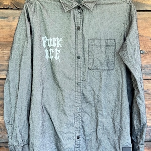 May include: A long-sleeved, button-down shirt in a heathered gray fabric. The shirt features a pointed collar, a single chest pocket on the left side, and a small square patch at the bottom hem. The text "FUCK ICE" is printed in white on the right chest.