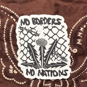 May include: A black and white sticker with the words "NO BORDERS NO NATIONS" in bold letters. The design includes a dandelion, barbed wire, and a chain-link fence, all in a graphic style. The sticker is on a brown patterned background.