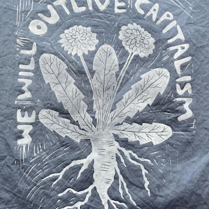 May include: A dark blue t-shirt with a white graphic print. The print features a stylized dandelion with the text "WE WILL OUTLIVE CAPITALISM" in a circular arrangement around the plant. The design has a linocut style.