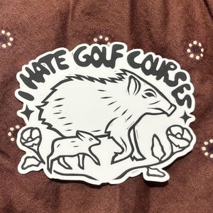 Peut inclure: Un autocollant blanc découpé avec des illustrations à l'encre noire représentant un pécari et son petit, entourés de fleurs et d'étoiles. Le texte "I HATE GOLF COURSES" est imprimé en lettres noires, grasses et arquées en haut du motif.
