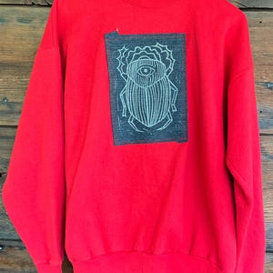 May include: A bright red long-sleeve sweatshirt featuring a dark gray rectangular patch on the chest. The patch displays a white line-art illustration of a beetle with a prominent eye on its back, stitched onto the fabric with red thread.