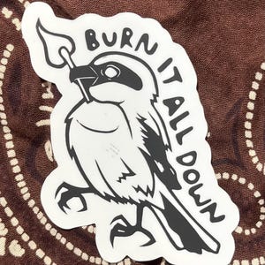 May include: A white sticker with a black and white illustration of a bird holding a lit match. The text "BURN IT ALL DOWN" is written around the bird in black, bold letters. The sticker is set against a brown patterned background.
