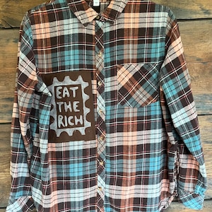 May include: A long-sleeve button-down flannel shirt featuring a plaid pattern in shades of brown, light blue, and peach. A brown rectangular patch on the front displays the hand-painted white text EAT THE RICH inside a scalloped border.