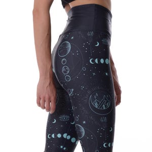 May include: Dark navy leggings with a celestial design featuring moons, stars, and mountain silhouettes in a light teal colour. The leggings have a high waistband and are worn with white and black trainers. The overall theme is activewear.
