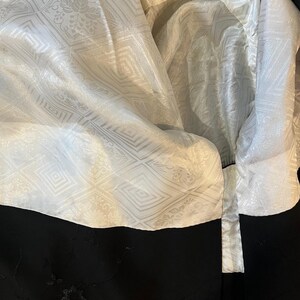 May include: A black garment featuring a white lining with a repeating geometric and floral jacquard pattern. The black exterior fabric displays delicate, dark embroidered floral details and subtle shimmering accents across the surface.