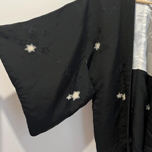 May include: A black kimono jacket featuring embroidered white floral motifs across the fabric. The garment has wide sleeves and a contrasting white inner lining with a subtle geometric pattern visible at the collar and front opening.