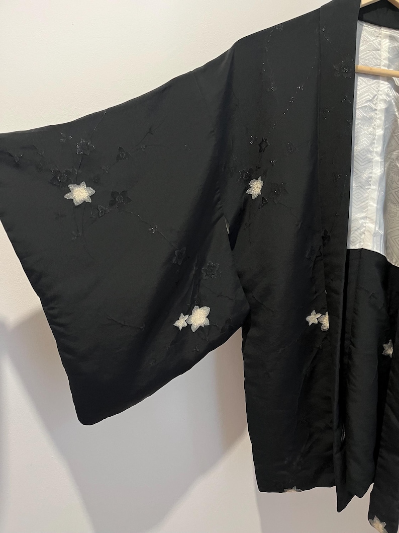 May include: A black kimono jacket featuring embroidered white floral motifs across the fabric. The garment has wide sleeves and a contrasting white inner lining with a subtle geometric pattern visible at the collar and front opening.