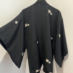 May include: A black kimono-style jacket featuring wide sleeves and a subtle, tonal floral pattern throughout. The garment is adorned with scattered, embroidered white cherry blossom motifs across the back and sleeves, hanging on a wooden hanger.