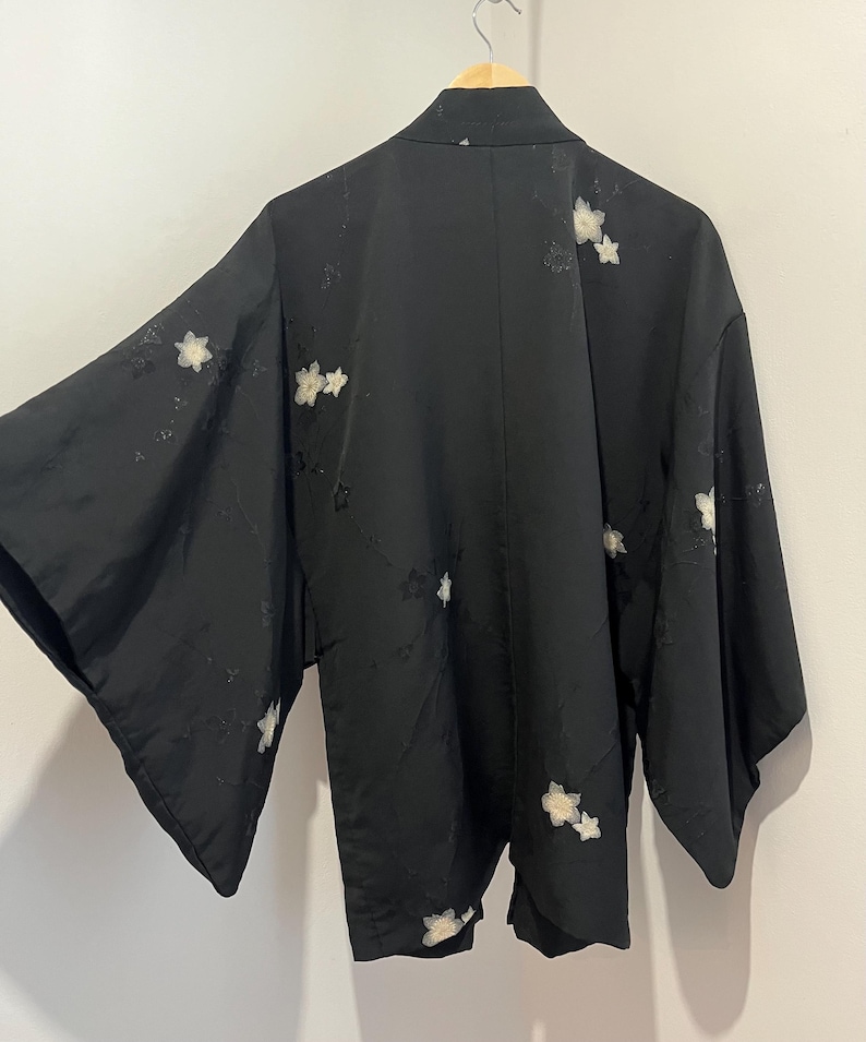 May include: A black kimono-style jacket featuring wide sleeves and a subtle, tonal floral pattern throughout. The garment is adorned with scattered, embroidered white cherry blossom motifs across the back and sleeves, hanging on a wooden hanger.