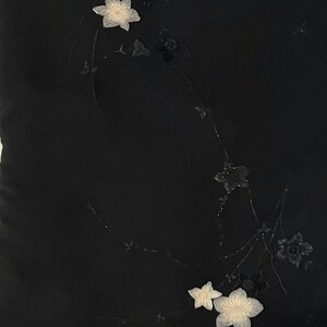 May include: A black fabric surface featuring delicate, embroidered floral motifs. The design includes several small, light-colored blossoms and thin, dark, trailing stems that extend across the dark background in a subtle, elegant arrangement.