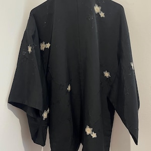 May include: A black haori jacket featuring a subtle tonal floral pattern and scattered embroidered white flower motifs. The garment has wide sleeves, a straight collar, and is displayed on a wooden hanger against a plain wall.