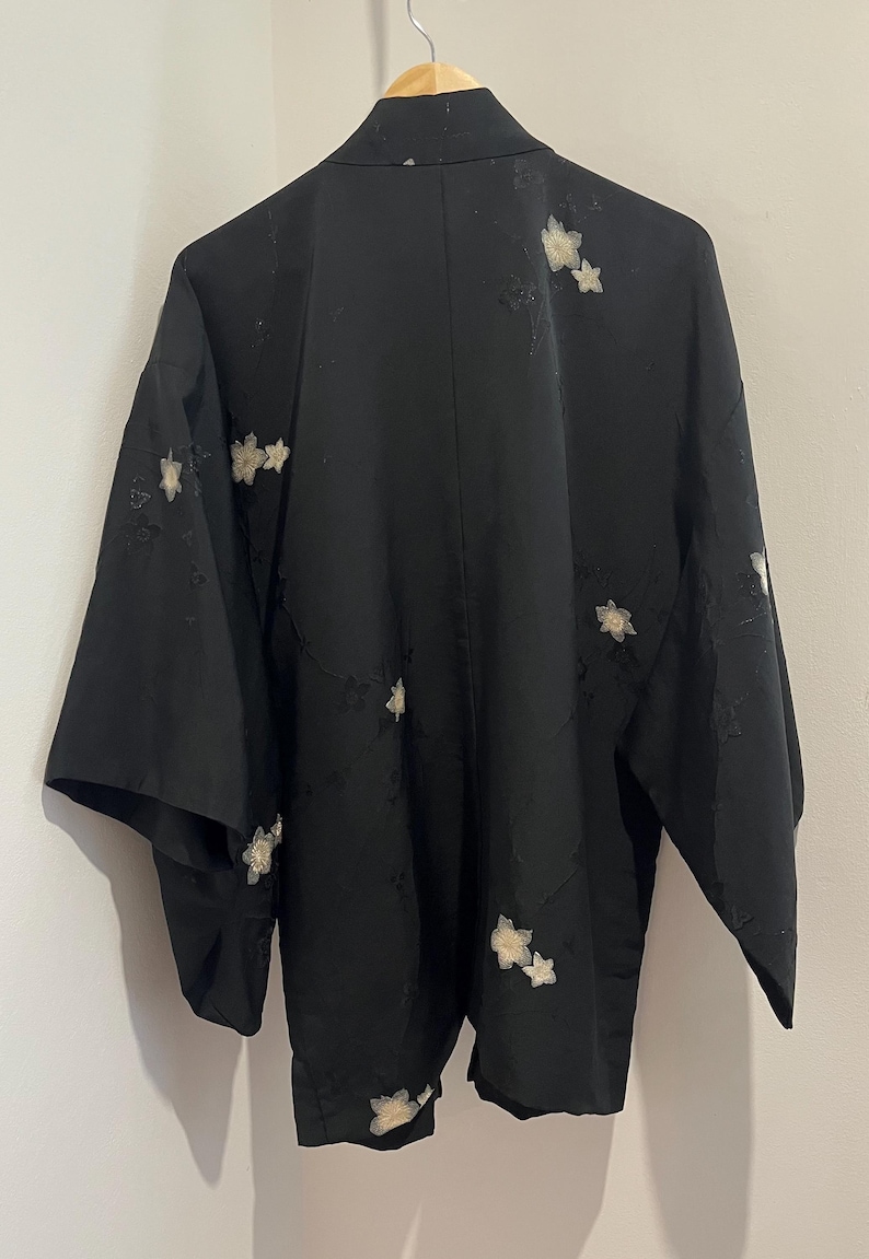 May include: A black haori jacket featuring a subtle tonal floral pattern and scattered embroidered white flower motifs. The garment has wide sleeves, a straight collar, and is displayed on a wooden hanger against a plain wall.