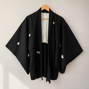 May include: A black haori jacket featuring wide sleeves and an open front design. The garment is decorated with small, white, star-shaped floral motifs scattered across the fabric. The interior lining is a contrasting white with a subtle pattern.
