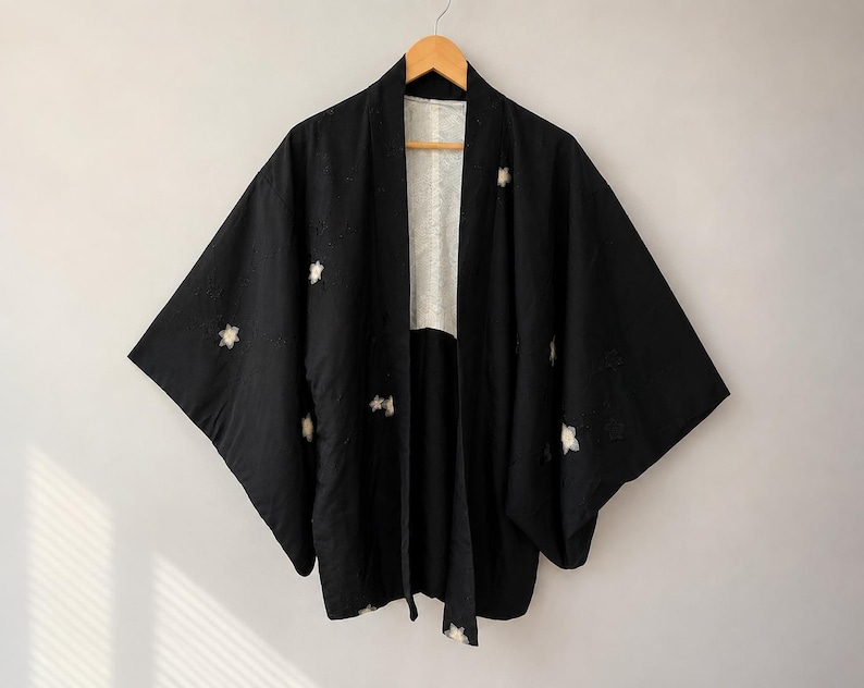 May include: A black haori jacket featuring wide sleeves and an open front design. The garment is decorated with small, white, star-shaped floral motifs scattered across the fabric. The interior lining is a contrasting white with a subtle pattern.