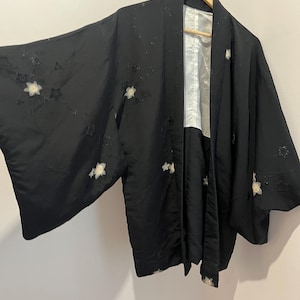 May include: A black haori jacket featuring wide sleeves and a white inner lining. The exterior fabric is adorned with scattered, embroidered white floral motifs and subtle tonal star-shaped patterns across the sleeves and front panels.