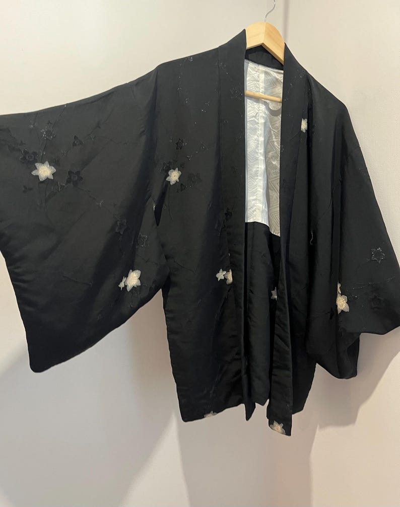 May include: A black haori jacket featuring wide sleeves and a white inner lining. The exterior fabric is adorned with scattered, embroidered white floral motifs and subtle tonal star-shaped patterns across the sleeves and front panels.