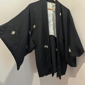 May include: A black haori jacket featuring wide sleeves and a white interior lining. The exterior fabric is decorated with scattered, embroidered floral motifs in white and black thread, creating a subtle textured pattern across the garment.