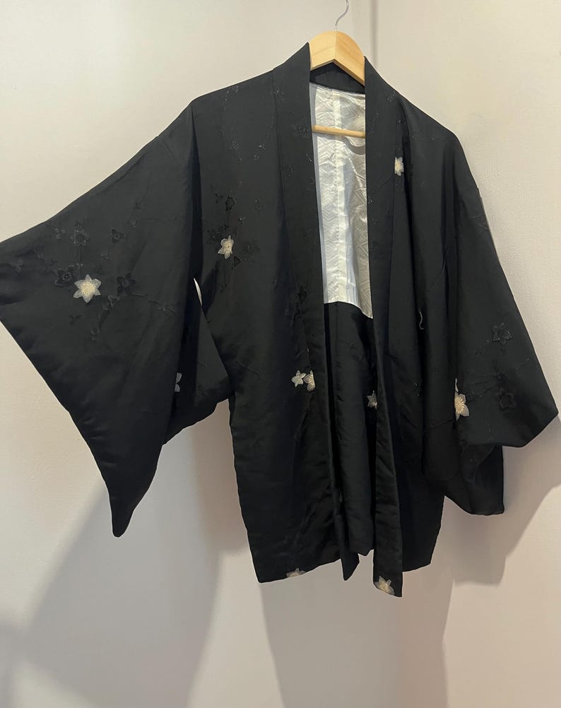 May include: A black haori jacket featuring wide sleeves and a white interior lining. The exterior fabric is decorated with scattered, embroidered floral motifs in white and black thread, creating a subtle textured pattern across the garment.