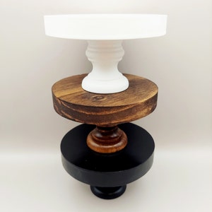 May include: A three-tiered display stand featuring a white top tier, a medium-toned brown wood middle tier, and a black bottom tier. Each circular platform is supported by a central pedestal base, creating a stacked structure with distinct colors.