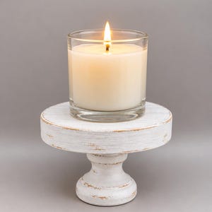 May include: A lit candle in a clear glass container resting on a small, round, white wooden pedestal stand with a distressed finish that reveals brown wood tones along the edges and base of the circular platform and the pedestal support.