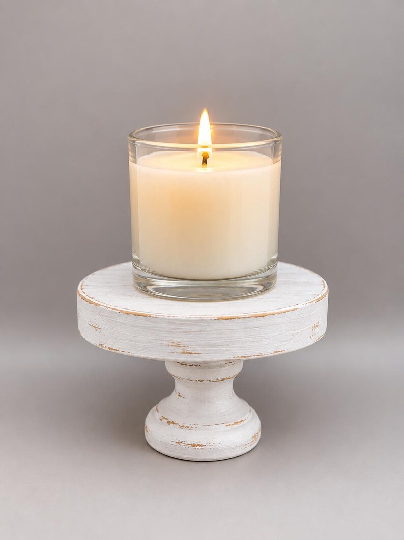 May include: A lit candle in a clear glass container resting on a small, round, white wooden pedestal stand with a distressed finish that reveals brown wood tones along the edges and base of the circular platform and the pedestal support.