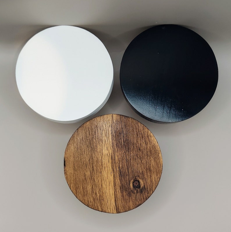 May include: Three circular lids arranged in a triangular formation. One lid is solid matte white, one lid is solid glossy black, and one lid features a natural brown wood grain finish with a visible knot detail on the surface of the wood.