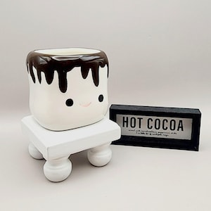 May include: A white ceramic mug shaped like a marshmallow, with a brown chocolate drizzle design and a smiling face. The mug sits on a small, white, square wooden stand with rounded legs. A black sign reads "HOT COCOA".
