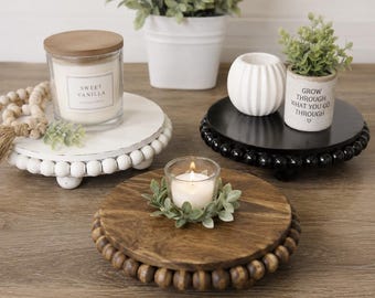 May include: Three round wooden risers with beaded edges in white, black, and natural wood finishes. The white riser holds a Sweet Vanilla candle, the black riser holds a white ribbed vase and a potted plant, and the natural wood riser holds a tea light.
