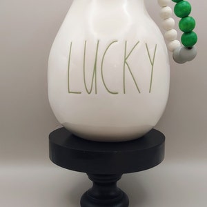 May include: A white ceramic pitcher with the word LUCKY printed in green lettering on the front. A decorative string of white, green, and gray wooden beads is attached to the handle. The pitcher rests on a black circular pedestal stand.