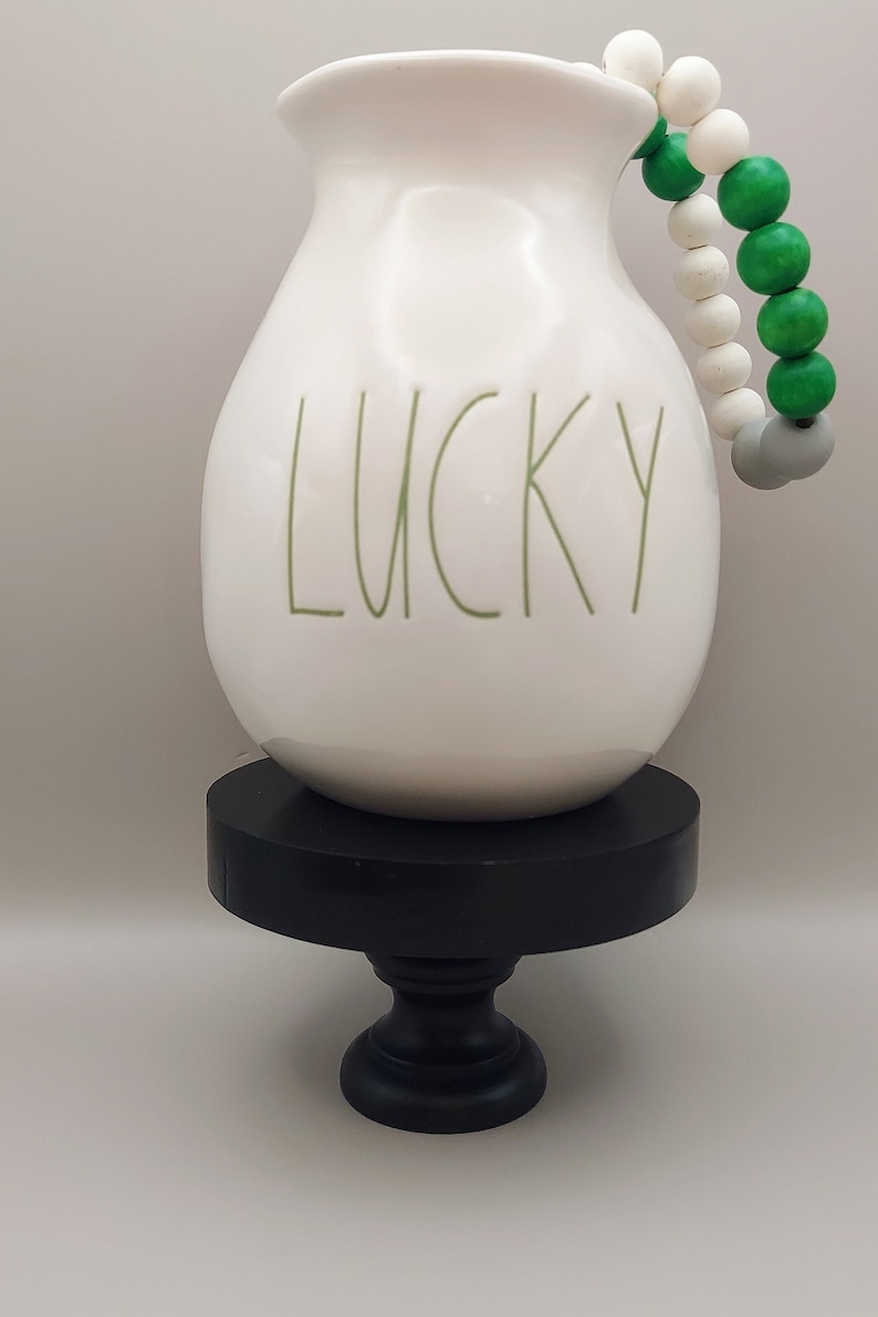 May include: A white ceramic pitcher with the word LUCKY printed in green lettering on the front. A decorative string of white, green, and gray wooden beads is attached to the handle. The pitcher rests on a black circular pedestal stand.