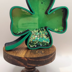 May include: A clover-shaped shaker ornament with a black and green frame, featuring a clear front panel filled with loose green beads, sequins, and glitter. The ornament is mounted on a small, round, dark brown wooden pedestal stand.