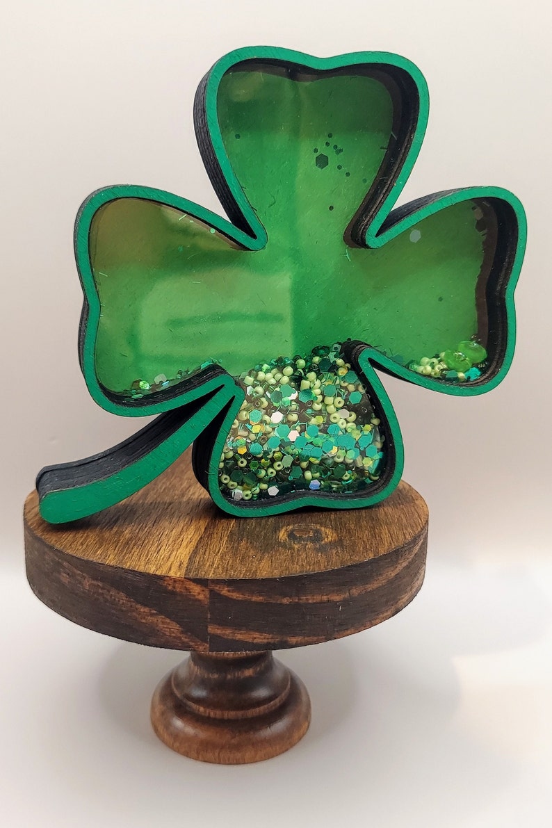 May include: A clover-shaped shaker ornament with a black and green frame, featuring a clear front panel filled with loose green beads, sequins, and glitter. The ornament is mounted on a small, round, dark brown wooden pedestal stand.