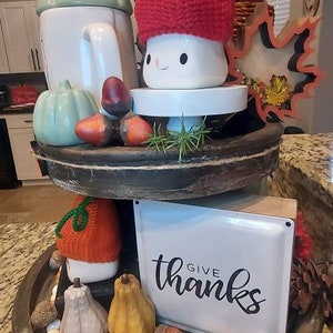 May include: A two-tiered wooden tray decorated for autumn. The top tier features a white mug with a red and tan knit hat, a light green pumpkin, and acorns. The bottom tier holds a white sign with the text "give thanks", pumpkins, and beads.