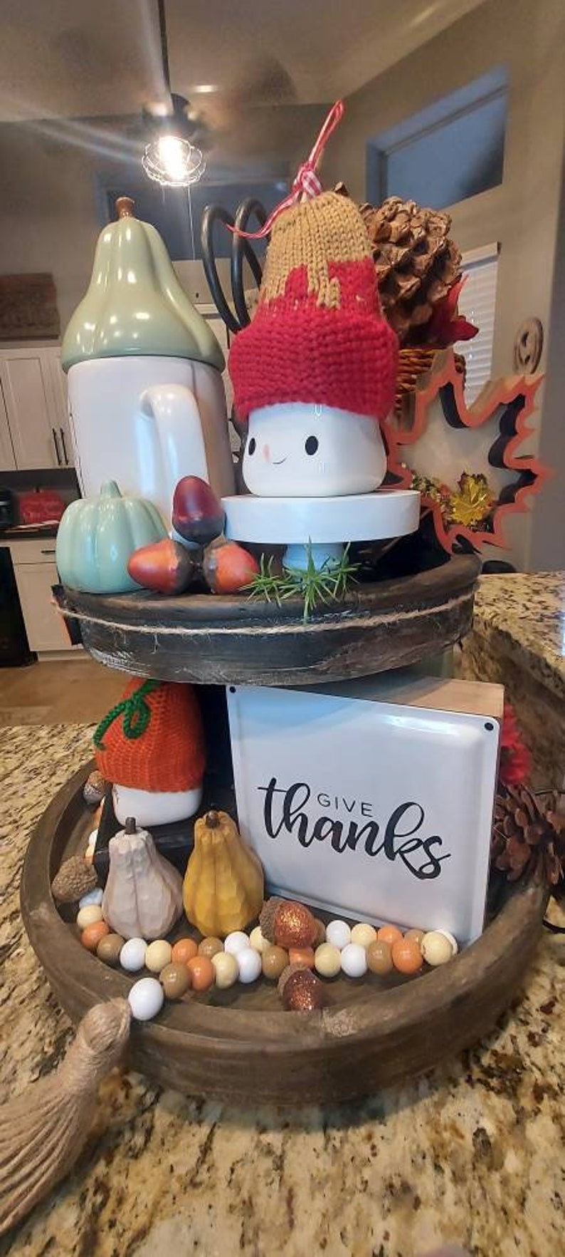 May include: A two-tiered wooden tray decorated for autumn. The top tier features a white mug with a red and tan knit hat, a light green pumpkin, and acorns. The bottom tier holds a white sign with the text "give thanks", pumpkins, and beads.