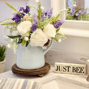 May include: A light blue ceramic watering can with the word "SHOWER" printed on the side, filled with a bouquet of white roses and purple lavender. The watering can is sitting on a wooden coaster and a small wooden sign with the text "JUST BEE" is in the foreground.