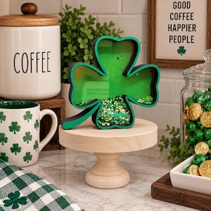 May include: A shamrock-shaped shadow box with a black frame and a green interior. The bottom section of the shamrock is filled with a mix of green, yellow, and white beads and sequins. The object is displayed on a light-colored wooden pedestal stand.
